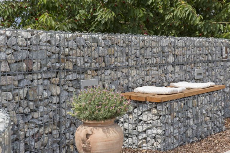 Eine mit Steinen gefüllte Gabionenwand mit einer Holzbank mit weißen Kissen neben einem großen Terrakotta-Topf mit blühenden Pflanzen, vor der Kulisse eines üppigen Gartens.