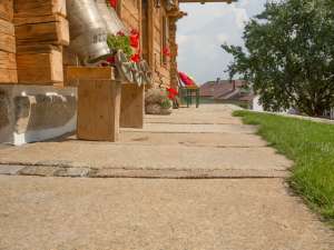 Eine Untersicht eines Steinpfads, der an einem Haus mit Holzverkleidung und leuchtend roten Blumen in einem Blumenkasten vorbeiführt und einen Eindruck von einer Vorstadtgegend vermittelt.