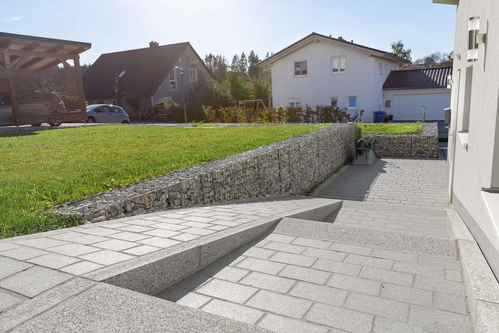 Eine sonnige Vorstadtszene mit einem hübsch angelegten Garten mit einer Gabionen-Stützmauer aus Stein und Stufen, die zu einer Rasenfläche führen, mit Häusern und einem geparkten Auto im Hintergrund.