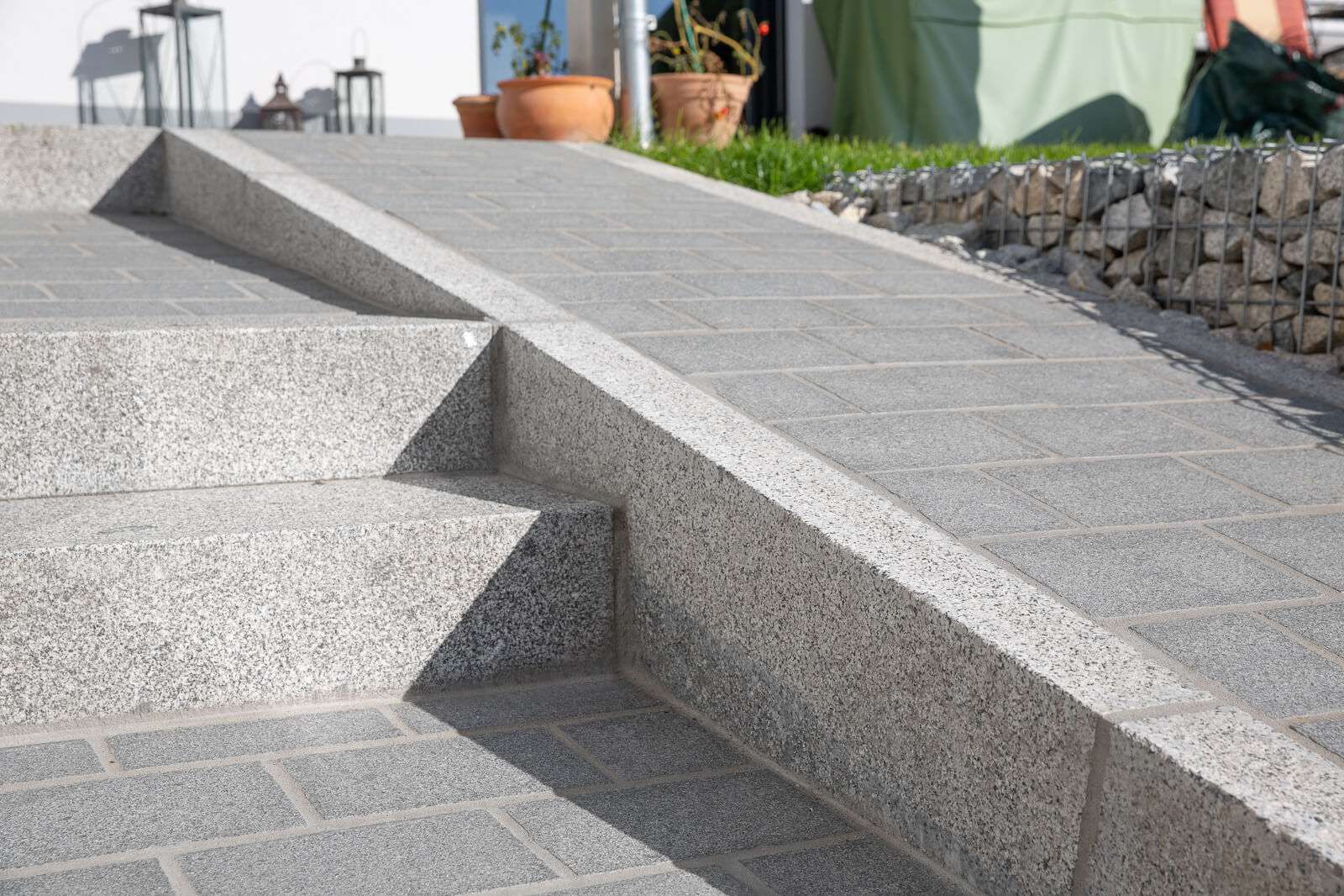 Nahaufnahme einer neu errichteten Außentreppe mit strukturierten grauen Fliesen und scharfen Winkeln und Kanten, die zu einem Terrassenbereich mit Pflanzen und Deko im Hintergrund führt.