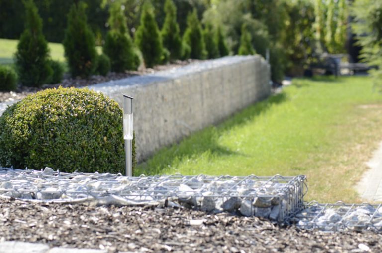 Eine fokussierte Ansicht einer solarbetriebenen Lampe auf einem Weg, mit verschwommenen Gabionenwänden und gepflegten Sträuchern im Hintergrund, in einem sonnigen Landschaftsgarten.