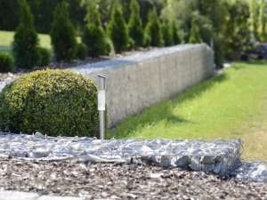 Eine fokussierte Ansicht einer solarbetriebenen Lampe auf einem Weg, mit verschwommenen Gabionenwänden und gepflegten Sträuchern im Hintergrund, in einem sonnigen Landschaftsgarten.