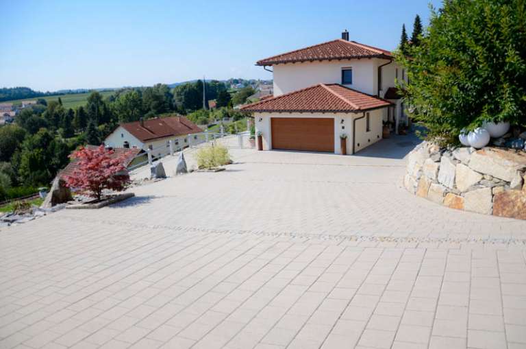 Eine geräumige Auffahrt mit hellen Pflastersteinen führt zu einem großen beigen Haus mit braunem Dach und Garagentor, umgeben von üppigem Grün und einer hügeligen Landschaft in der Ferne.