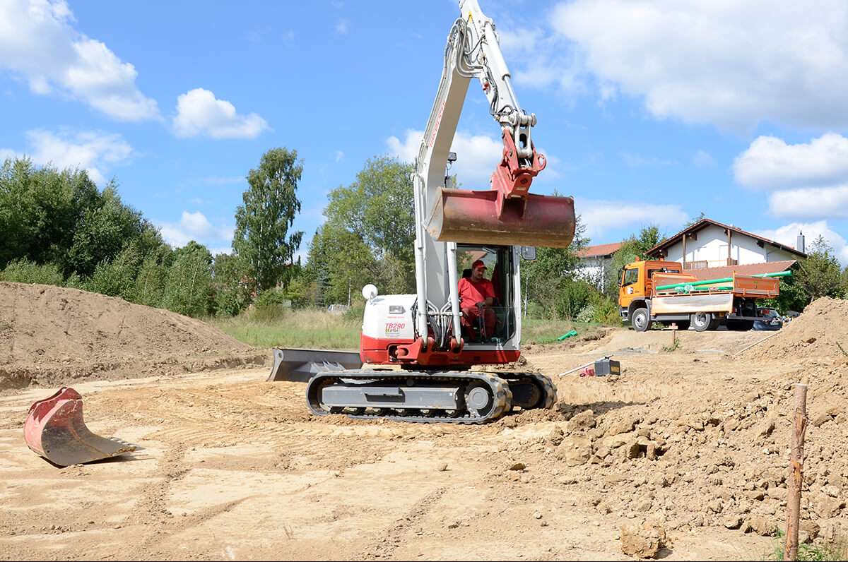 Eine Baustelle mit einem Arbeiter, der einen rot-weißen Bagger bedient. Der Bagger bewegt Erde und im Hintergrund ist ein Muldenkipper geparkt. In der Umgebung liegen Erdhaufen und in der Ferne sind unter einem teilweise bewölkten Himmel Bäume und ein Haus zu sehen.